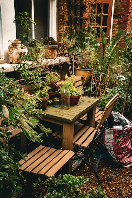 A cozy urban garden outdoor space featuring a weathered wooden table surrounded by matching wooden slatted chairs and a black metal bench, all set on a gravel surface. The garden is densely populated